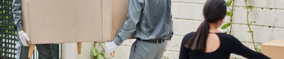 Two professional movers in uniform carry a beige sofa into a residence while a woman gestures toward a stack of cardboard boxes, illustrating the reliable services of movers Newark NJ.