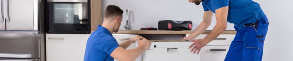 Two professional movers in blue uniforms are installing a dishwasher in a modern kitchen, with tools placed on the countertop representing skilled appliance handling by movers NJ.