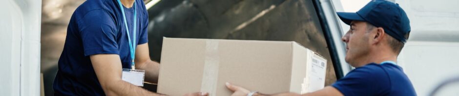 An image of two professional movers dressed in navy-blue uniforms and caps, working together to unload a large cardboard box from the back of a white moving van—highlighting a seamless “movers and packers NJ” operation