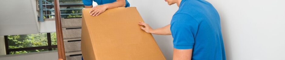 Two movers in blue uniforms and matching caps carefully carry a large cardboard box down a stairwell, using teamwork to navigate the steps in what appears to be a residential building in Newark, NJ.