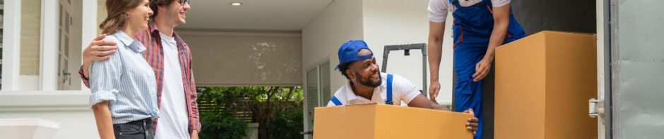 A smiling young couple standing outside their home as two professional movers and packers NJ staff unload large cardboard boxes from a moving truck at the driveway